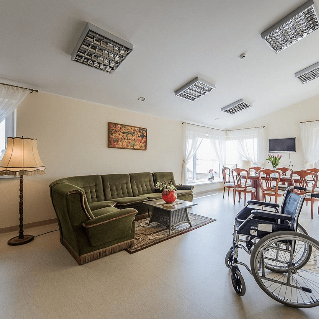 Erdvi ir šviesi globos namų bendroji erdvė su sofa, kavos staliuku, valgomojo zona ir neįgaliojo vežimėliu