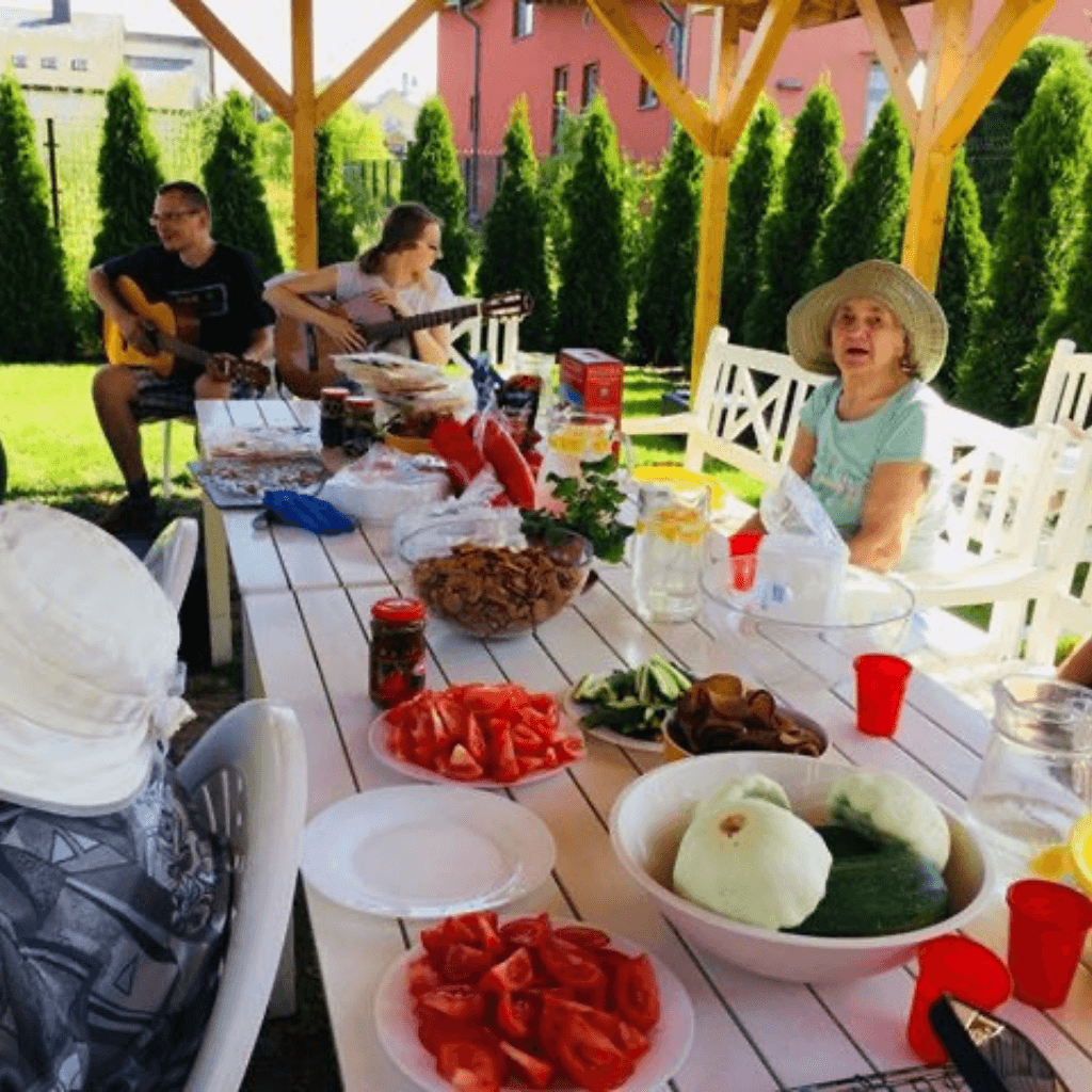 Globos namų gyventojai lauke pavėsinėje leidžia laiką prie bendro stalo, klausosi gitaros muzikos ir vaišinasi