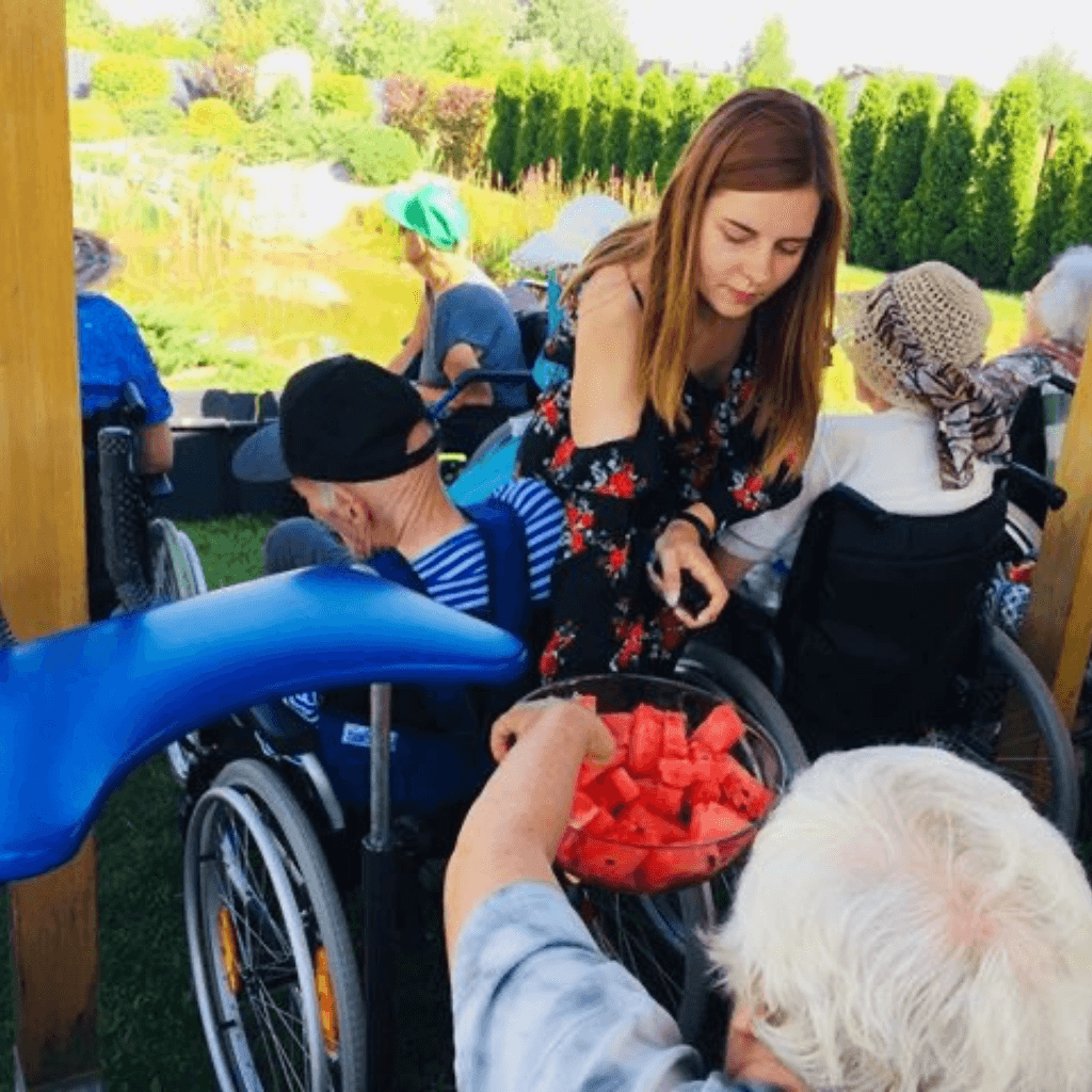 Globos namų gyventojai lauke su darbuotoja mėgaujasi arbūzu sodo teritorijoje