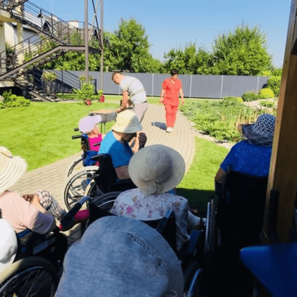 Globos namų gyventojai vežimėliuose leidžia laiką lauke, darbuotojai padeda pasivaikščiojimo metu