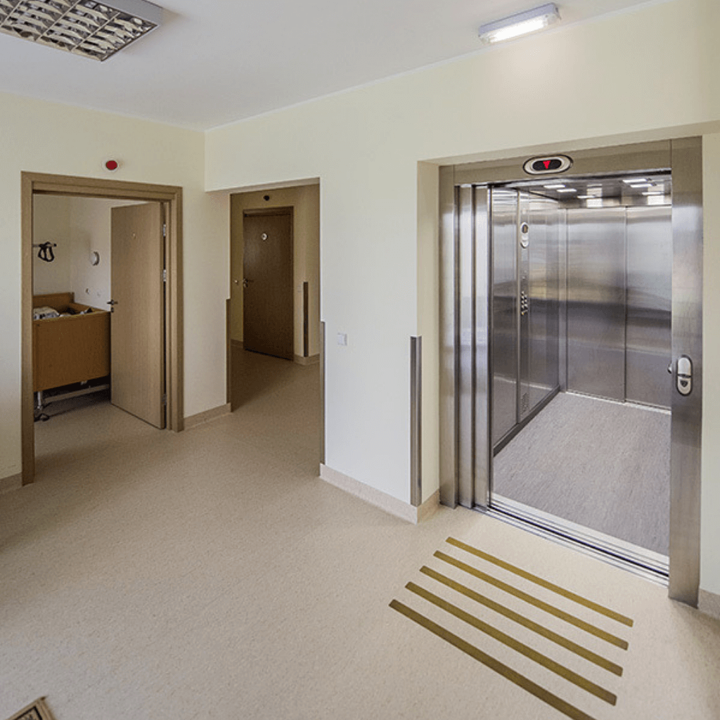 Globos namų koridorius su liftu ir erdviu praėjimu, pritaikytu gyventojų judėjimui