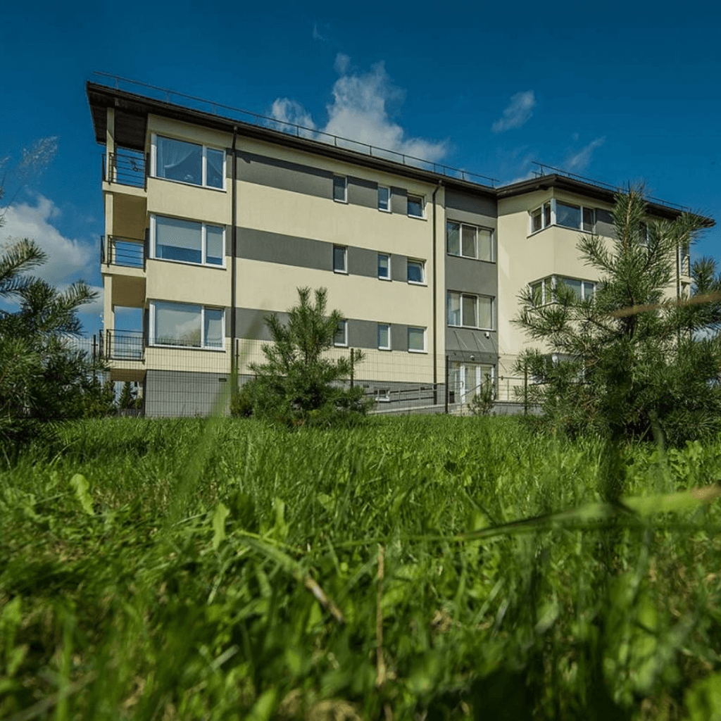 Modernus daugiabutis su balkonais, apsuptas žalios vejos ir medžių, ramioje gyvenamojoje aplinkoje.