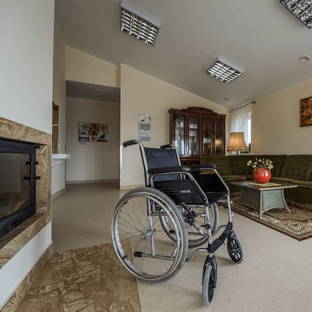 Neįgaliojo vežimėlis jaukioje senelių namų svetainėje su židiniu, sofa ir medine vitrina fone