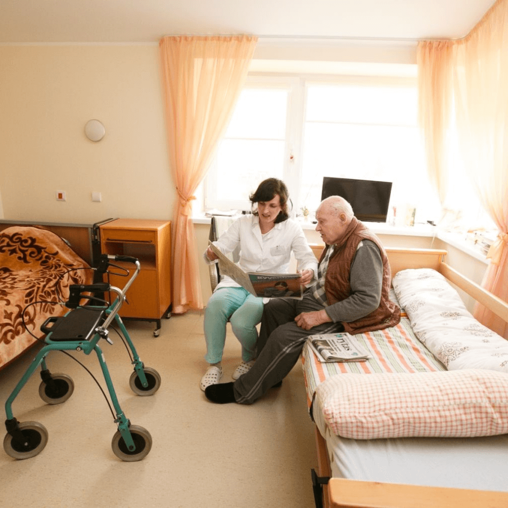 Slaugytoja senelių namuose kartu su senjoru skaito laikraštį kambaryje, šalia stovi vaikštynė
