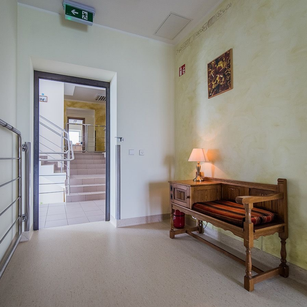 Šviesus pastato koridorius su laiptais, mediniu suoliuku ir jaukia, tvarkinga aplinka